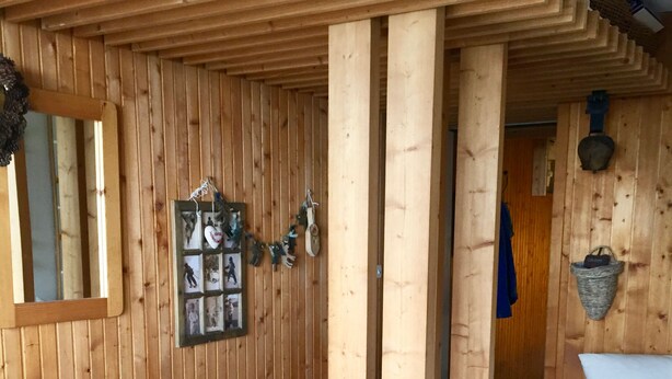 Una stanza in legno con un letto, un tavolino, una poltrona e uno scaffale a parete con fotografie e un cesto.