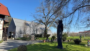 A building with a metal roof, a statue in the foreground, and a well-maintained garden with potted plants. - Vacation apartment at Obstgut Willmann, quiet location with a view of Lake Constance (Kressbronn)