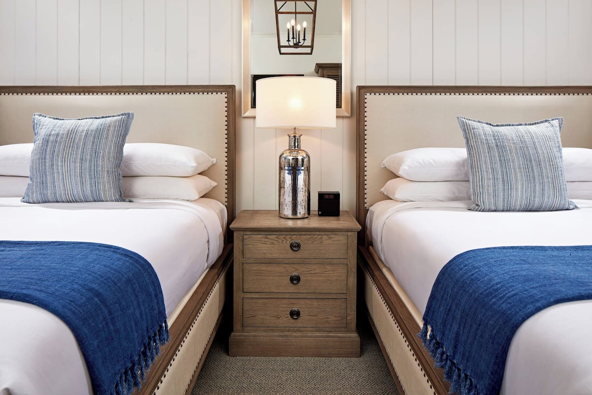 Une chambre d’hôtel avec deux lits, chacun agrémenté d’un oreiller à rayures bleues, une table de chevet en bois avec une lampe et un miroir au-dessus de la table de chevet.