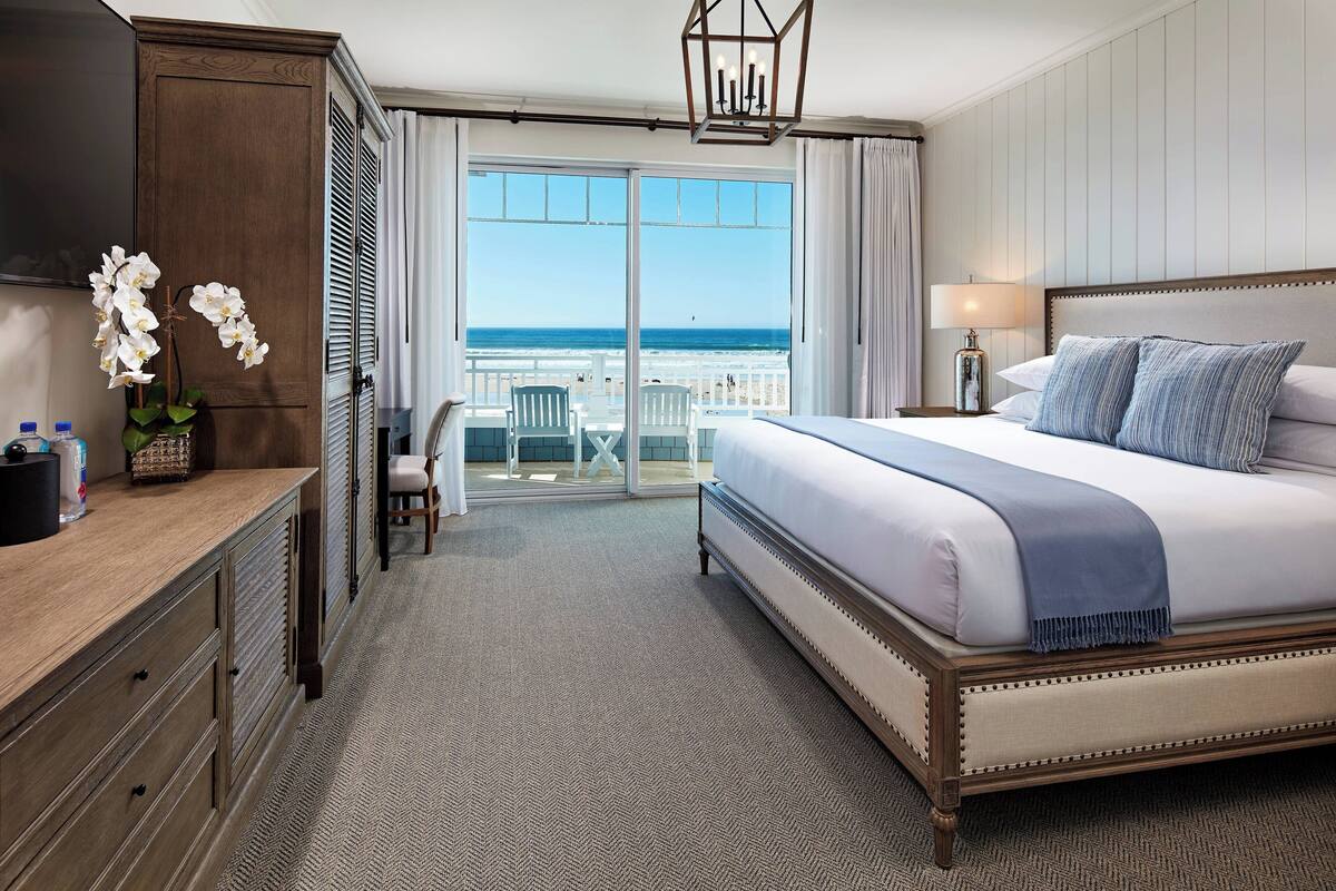 Une chambre d’hôtel avec un grand lit, un bureau avec une lampe, une chaise et une vue sur la plage, visible à travers les portes coulissantes.