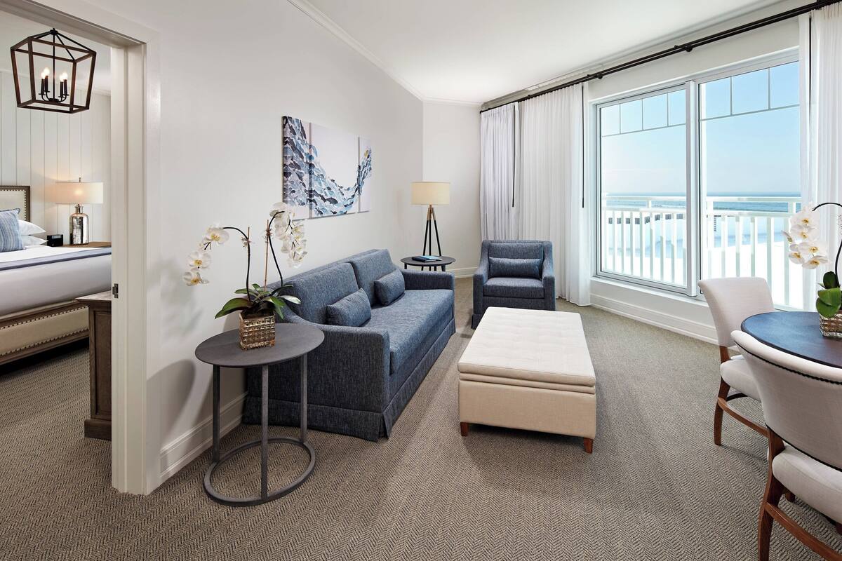 Une chambre d’hôtel avec un lit, un canapé bleu, un fauteuil gris, une table ronde et une vue sur l’océan.