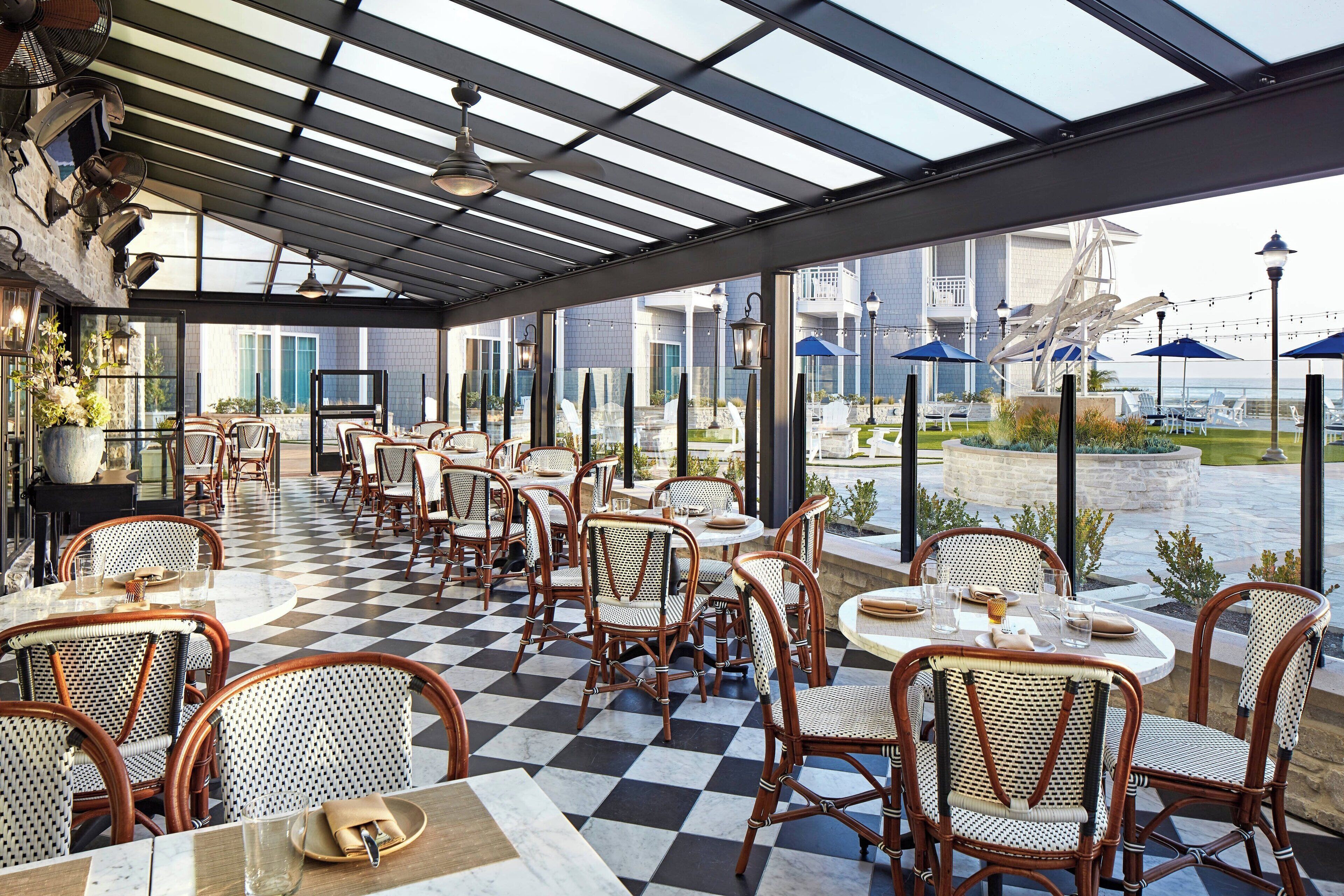 A covered outdoor seating area with checkered flooring, wicker chairs, and tables.