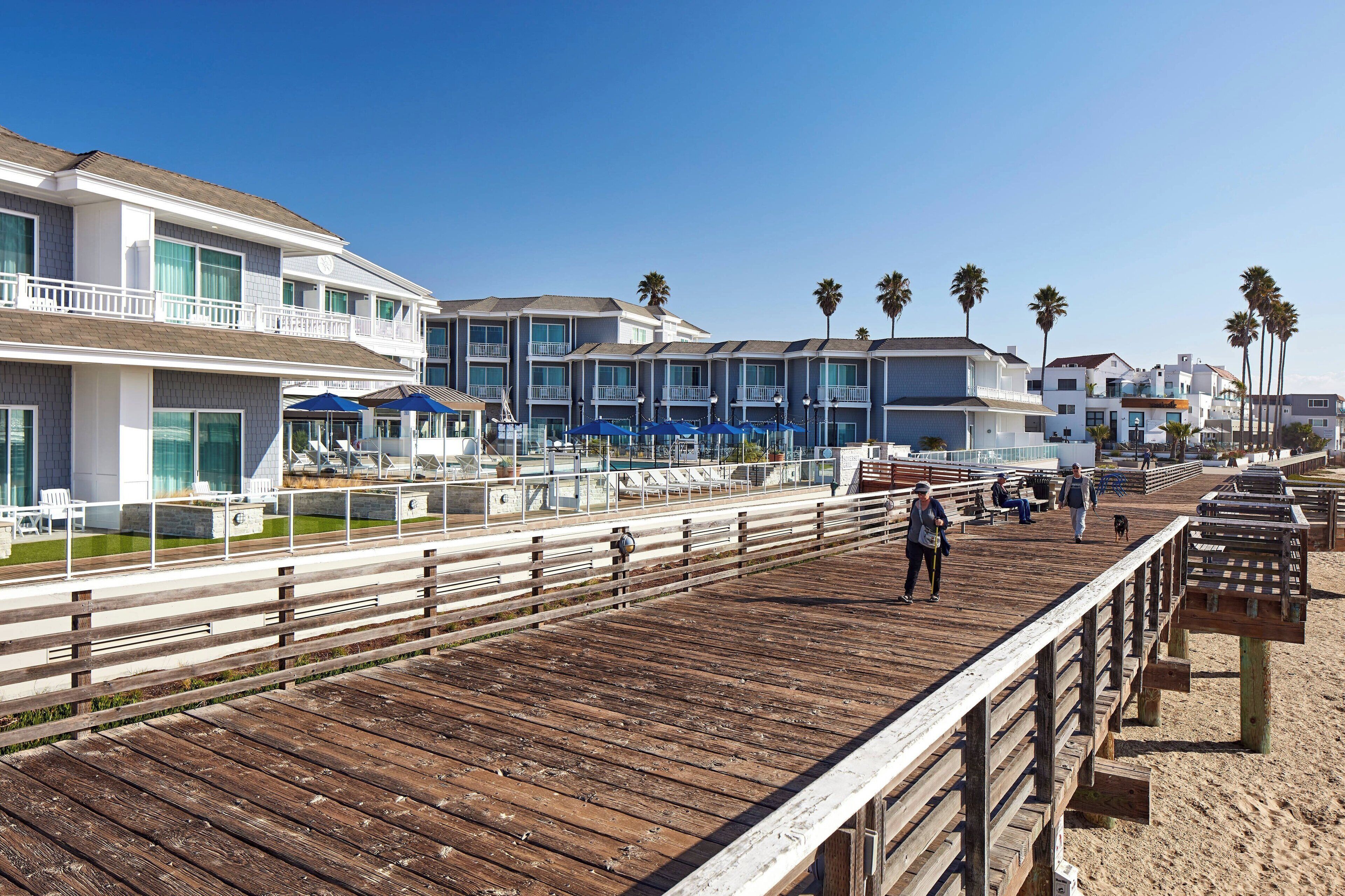 Une promenade en bois dotée d’une rambarde, qui mène à un hôtel en bord de mer, avec de nombreux balcons et un espace piscine.