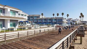 A wooden boardwalk with a railing, leading to a beachfront hotel with multiple balconies and a pool area.
