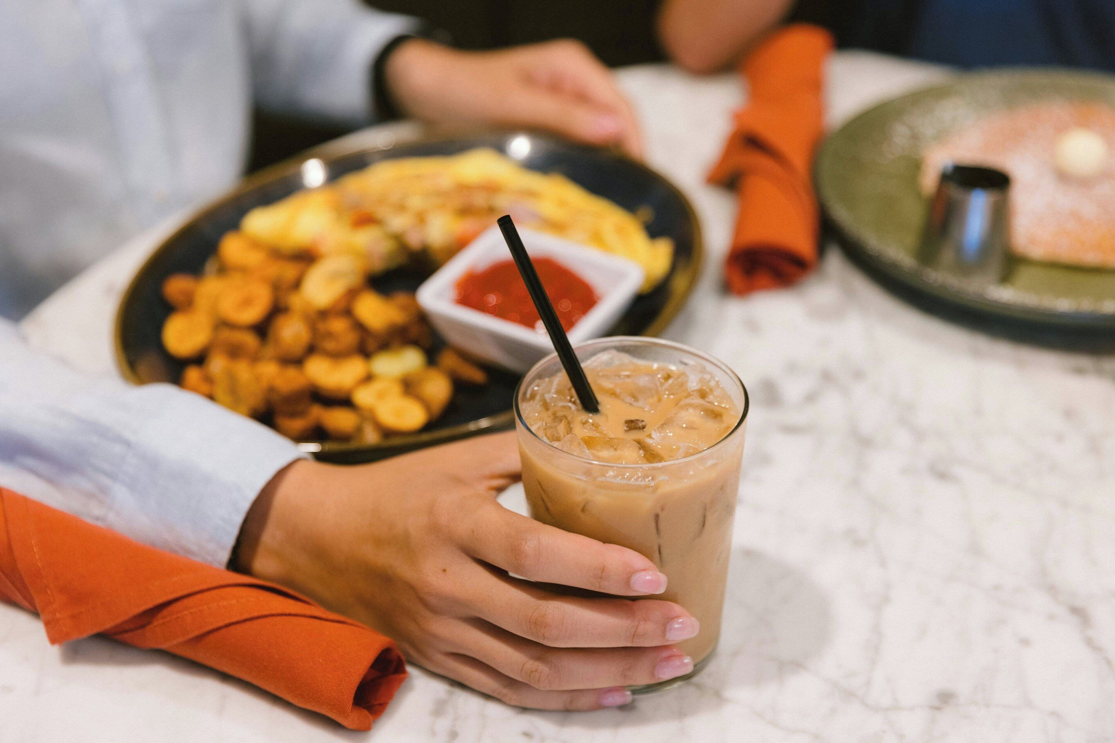 A person holding a glass of iced coffee with a straw, a plate of food, and a napkin on a table.