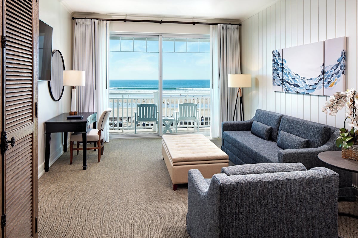 Une chambre d’hôtel avec un balcon, un canapé, un fauteuil, une petite table et un bureau.
