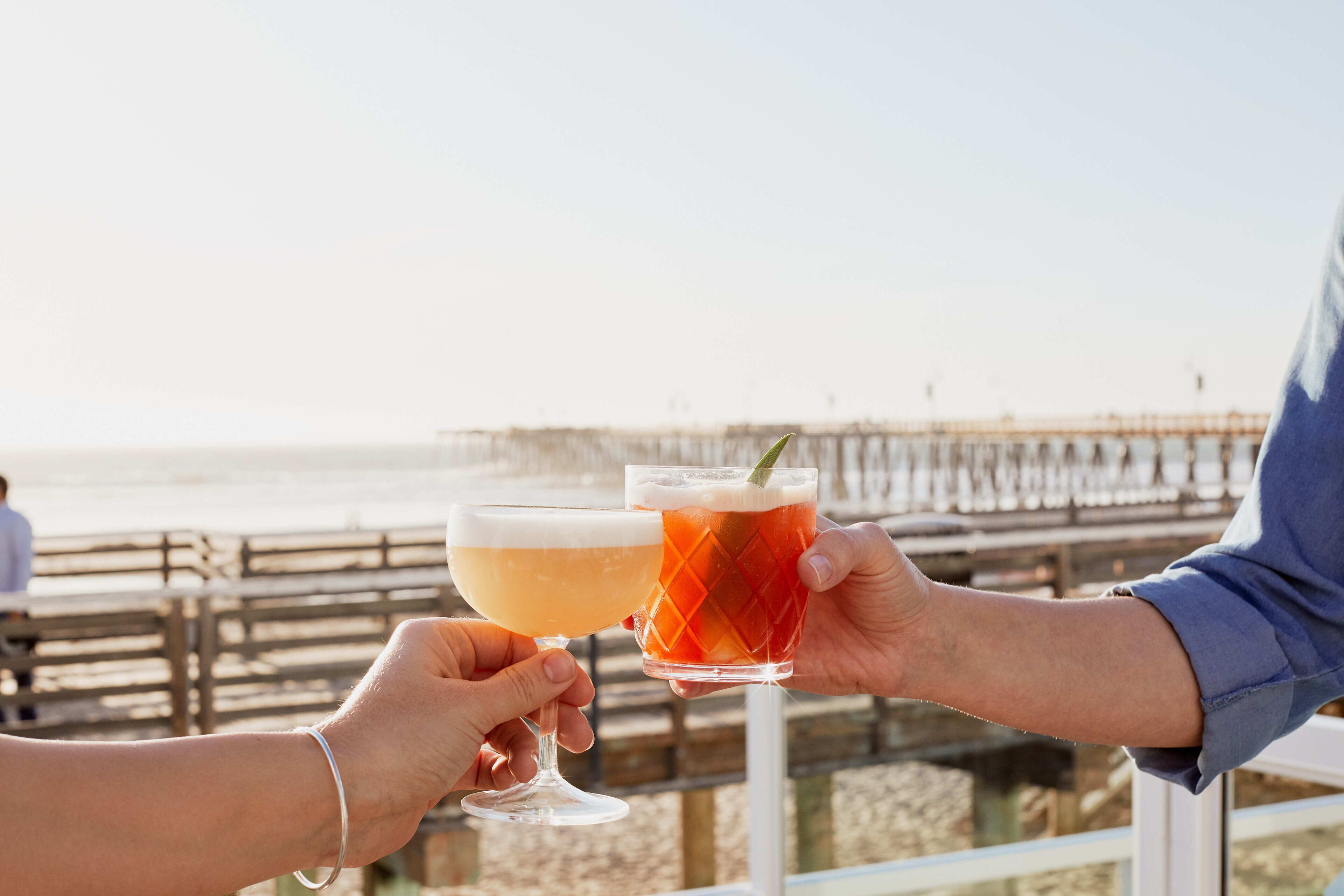 Two people holding cocktail glasses with a beach and pier in the background.