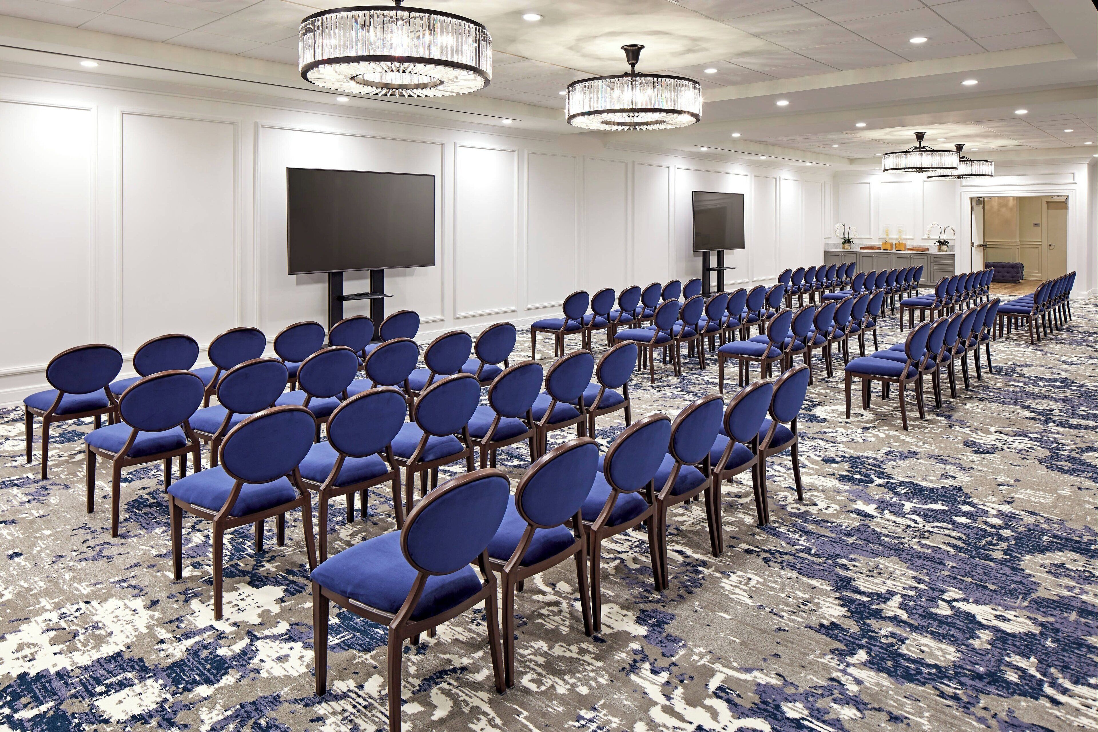 A conference room with rows of blue chairs, a large flat-screen TV, and chandeliers.