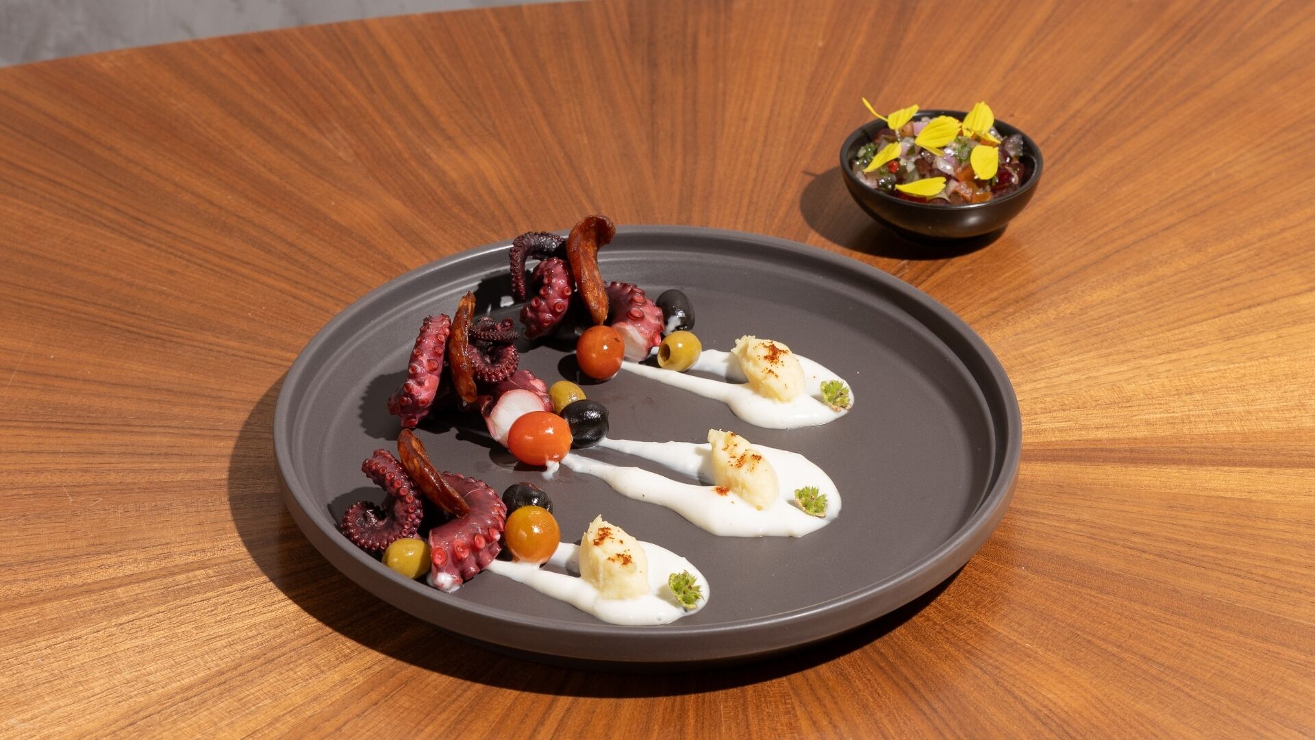 A plate of octopus tentacles, cherry tomatoes, olives, and a small bowl of yellow fruit on a wooden table.