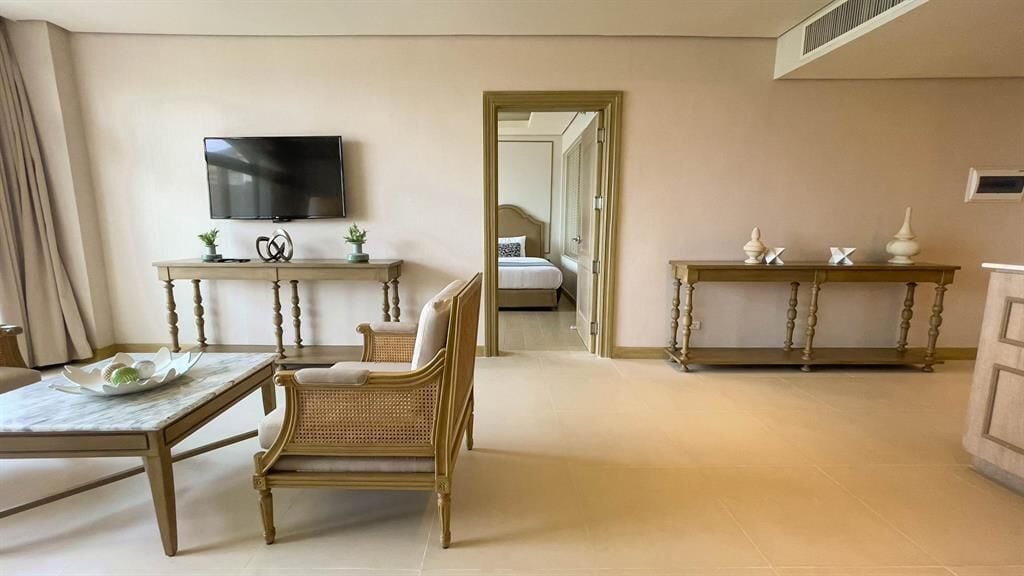 Une chambre d’hôtel équipée d’une télévision, d’une table à manger, d’un fauteuil en osier et de deux petites tables d’appoint ornées d’objets décoratifs.