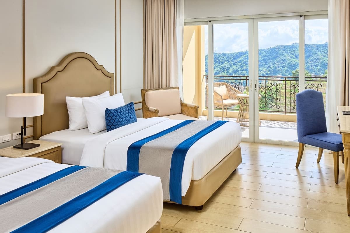 A hotel room with two beds, a desk, a chair, and a balcony with a view of the mountains.