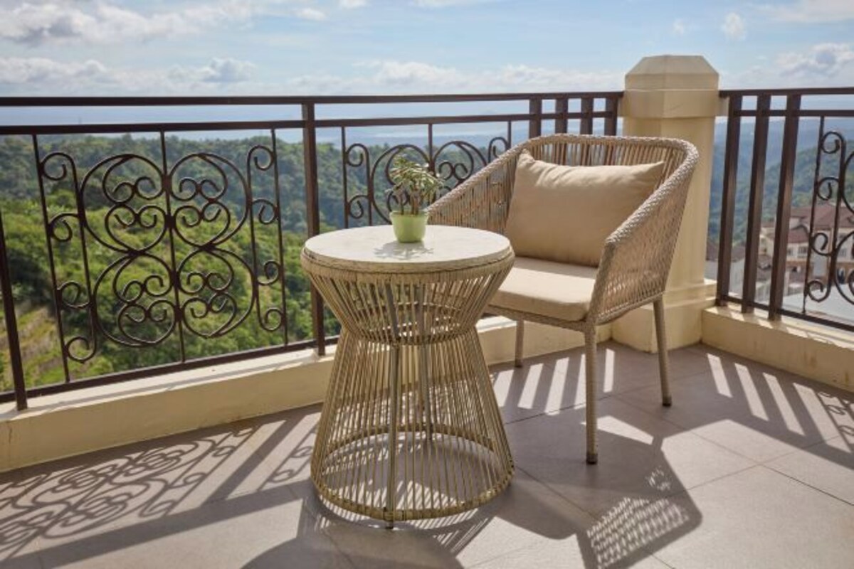 Un balcón con una silla de mimbre, una mesita redonda, una planta en maceta y vistas a un paisaje montañoso.
