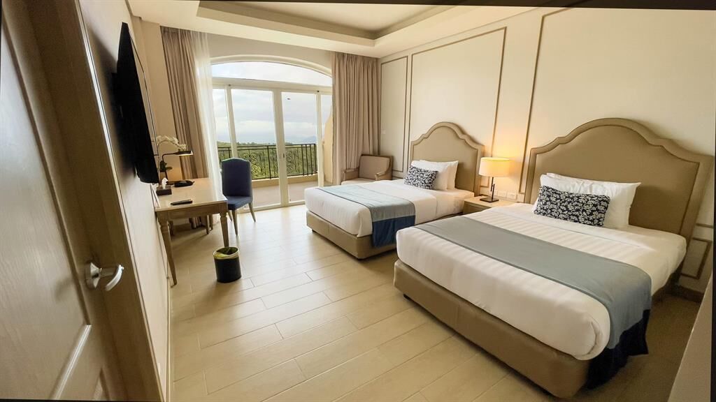 A hotel room with two beds, a desk, a chair, a TV, and a balcony with a view.