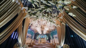 Un espacio interior con escenario, cortinas decorativas, adornos florales y mesas dispuestas para un evento.