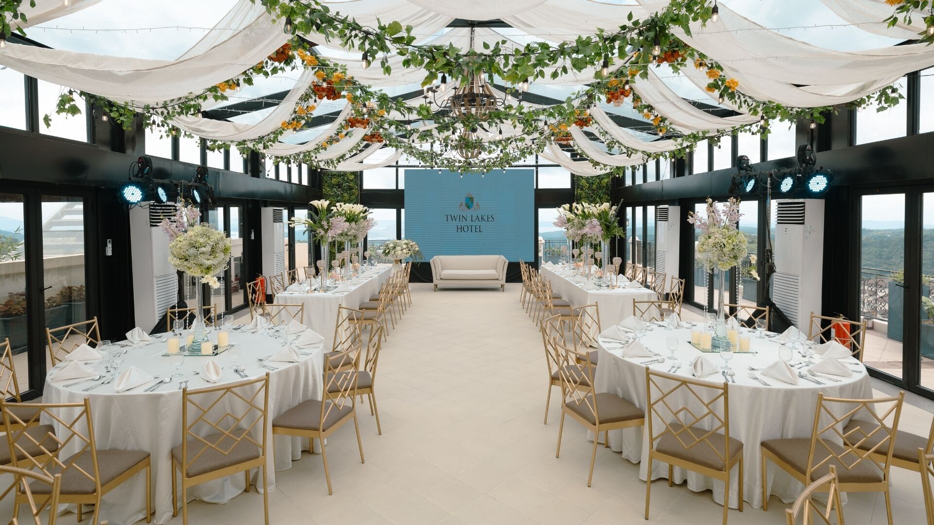 Montaje formal para comedor con mesas redondas, sillas doradas y manteles blancos, con un sofá y un cartel que dice "Twin Lakes Hotel" en el centro.