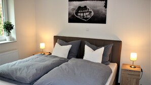 A bedroom with a bed, two bedside lamps, a nightstand, a potted plant, and a framed picture on the wall. - Apartment / Fitter Eal at the Thülsfelder dam (Friesoythe)