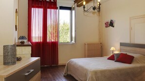 A bedroom with a bed, a nightstand, a dresser, a window with red curtains, a radiator, and a wall-mounted light fixture.