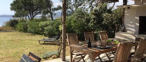 Un espace de restauration en plein air avec une table et des chaises en bois, une pergola au-dessus et une vue sur la mer et les arbres en arrière-plan.