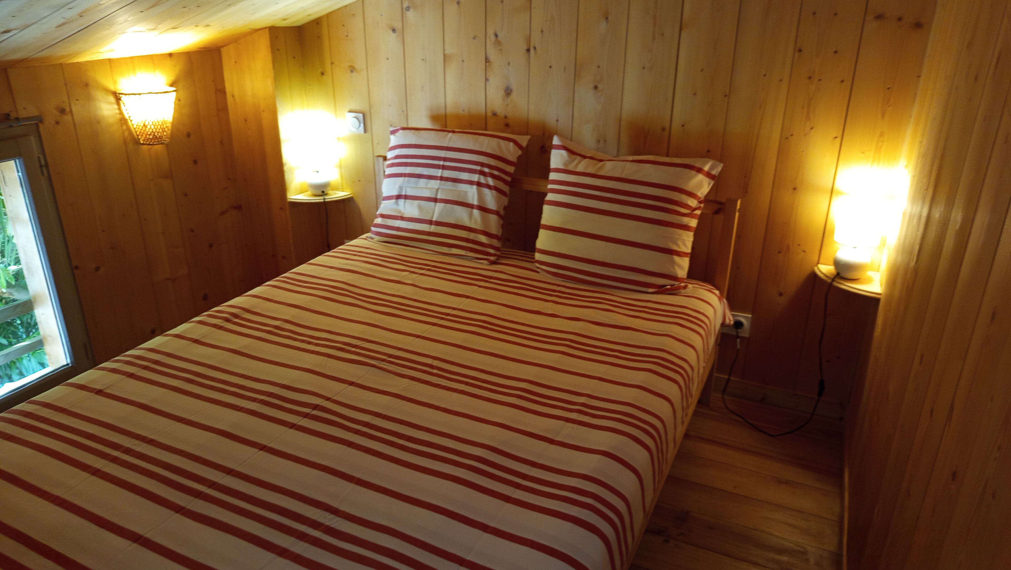 A wooden room with a bed, striped bedding, and two wall-mounted lamps.