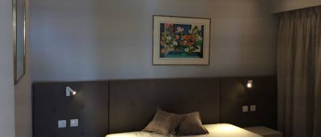 A hotel room with a large bed, two bedside tables, a nightstand, a framed floral artwork on the wall, and a window with curtains.