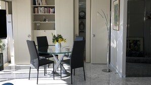 Een moderne eethoek met een glazen tafel en stoelen, een boekenkast vol boeken en decoratieve objecten, en een marmeren vloer.