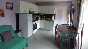 Cocina compacta con gabinetes blancos, un refrigerador, microondas y cafetera. Hay un comedor con mesa y sillas, y un sofá verde con un cojín.