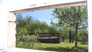 A view through a window of a room with a wooden floor, a stone pillar, and a view of a backyard with a wooden fence, trees, and grass. - *** Perigord cottage with pool (Archignac)