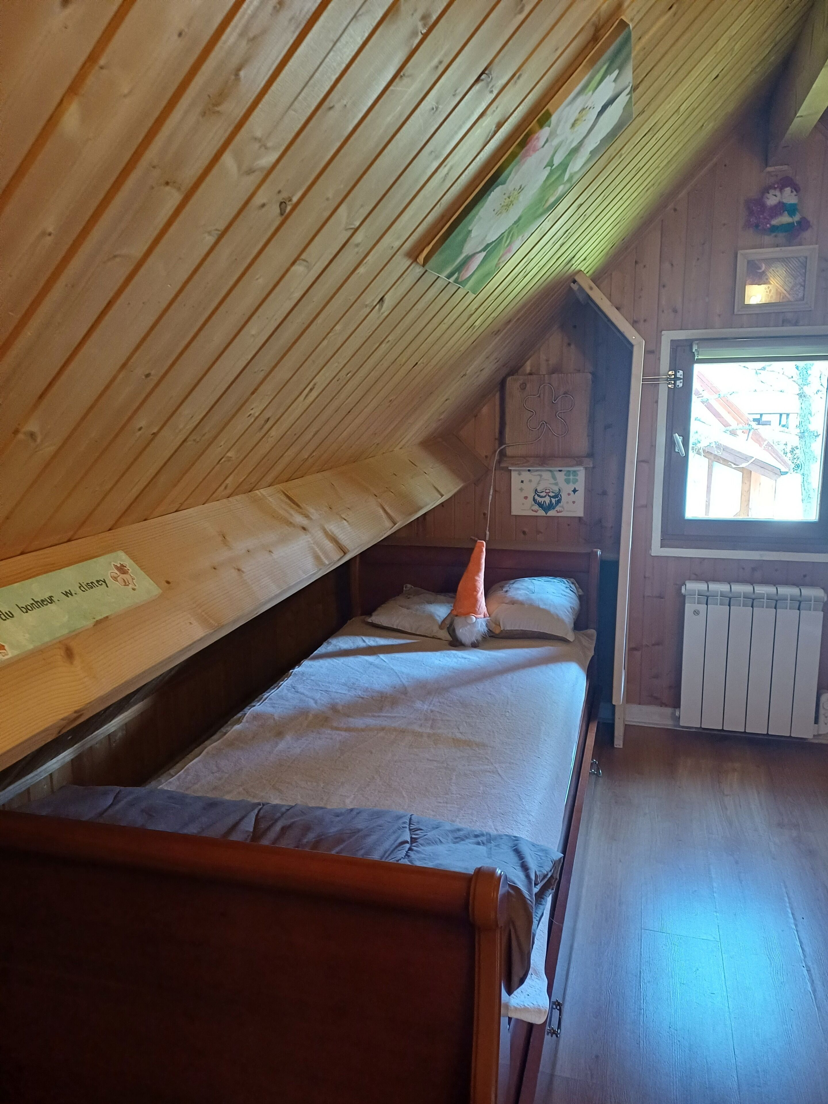 Een houten kamer met een schuine kap, een bed met een witte matras en een oranje kussen, een raam met uitzicht op een gebouw, en een radiator aan de muur.
