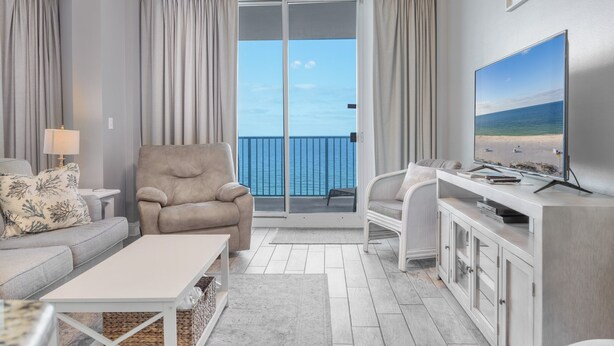 Una sala moderna con sofá, sillón y televisor, un balcón con cortinas y vista al océano.