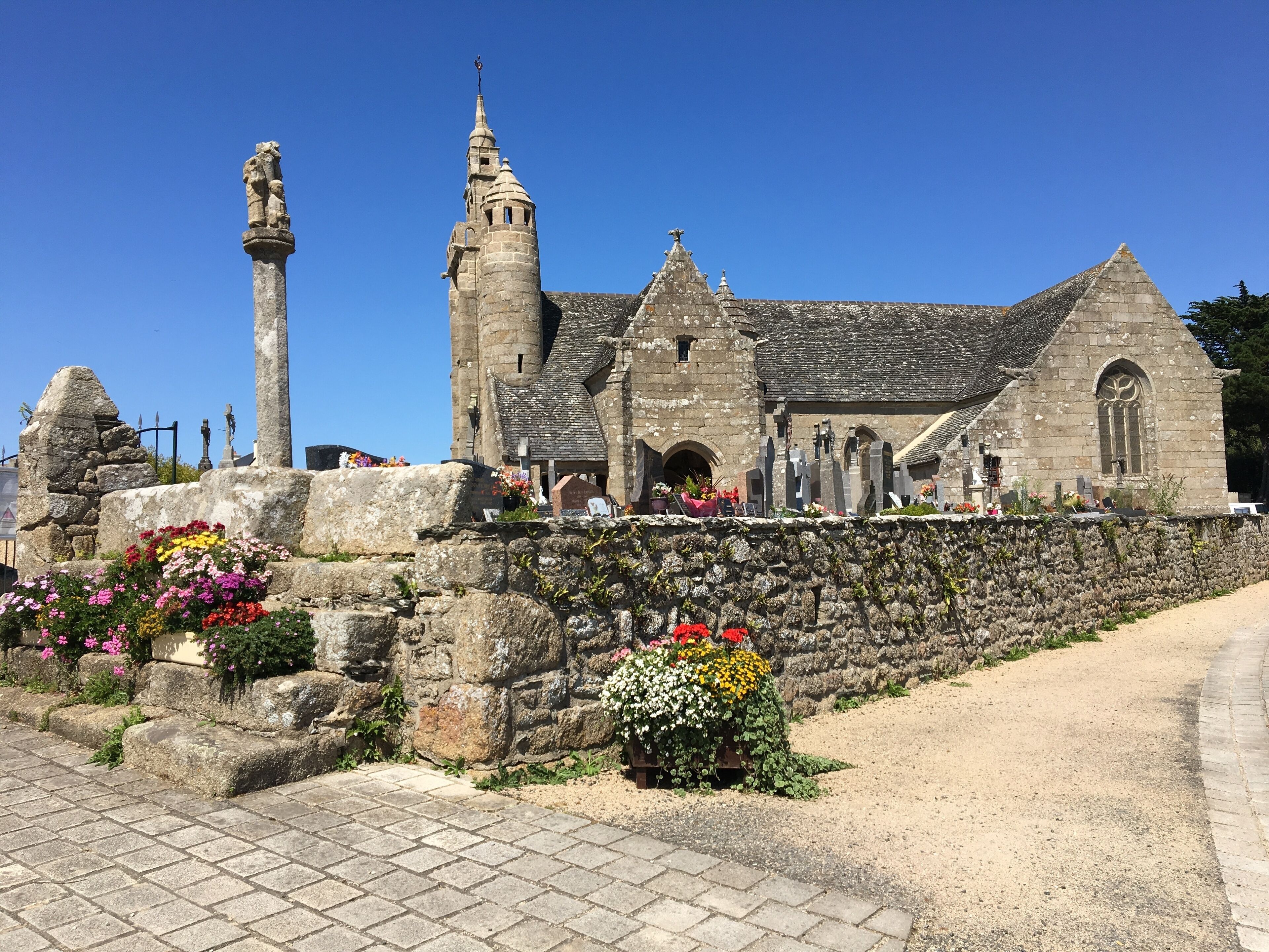 A historic stone building with a tall tower and a smaller spire, surrounded by a stone wall and flower beds.