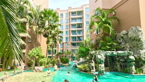 Zona de piscina con varios niveles de balcones, rodeada de palmeras y una formación rocosa.