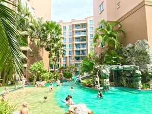 Un espace piscine doté de plusieurs niveaux de balcons, entouré de palmiers et agrémenté d’un élément décoratif en roche.