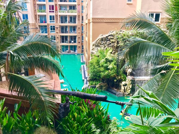 A pool with a waterfall, surrounded by lush greenery and palm trees, with a view of a multi-story building in the background. - Atlantis Condo Resort (Pattaya)