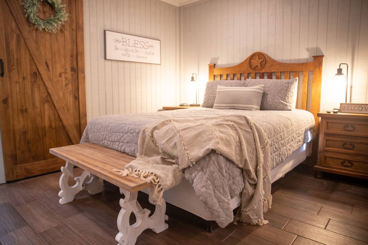 A bedroom with a wooden bed, a bench at the foot of the bed, a wooden dresser, and a wall with a 'Bless' sign.