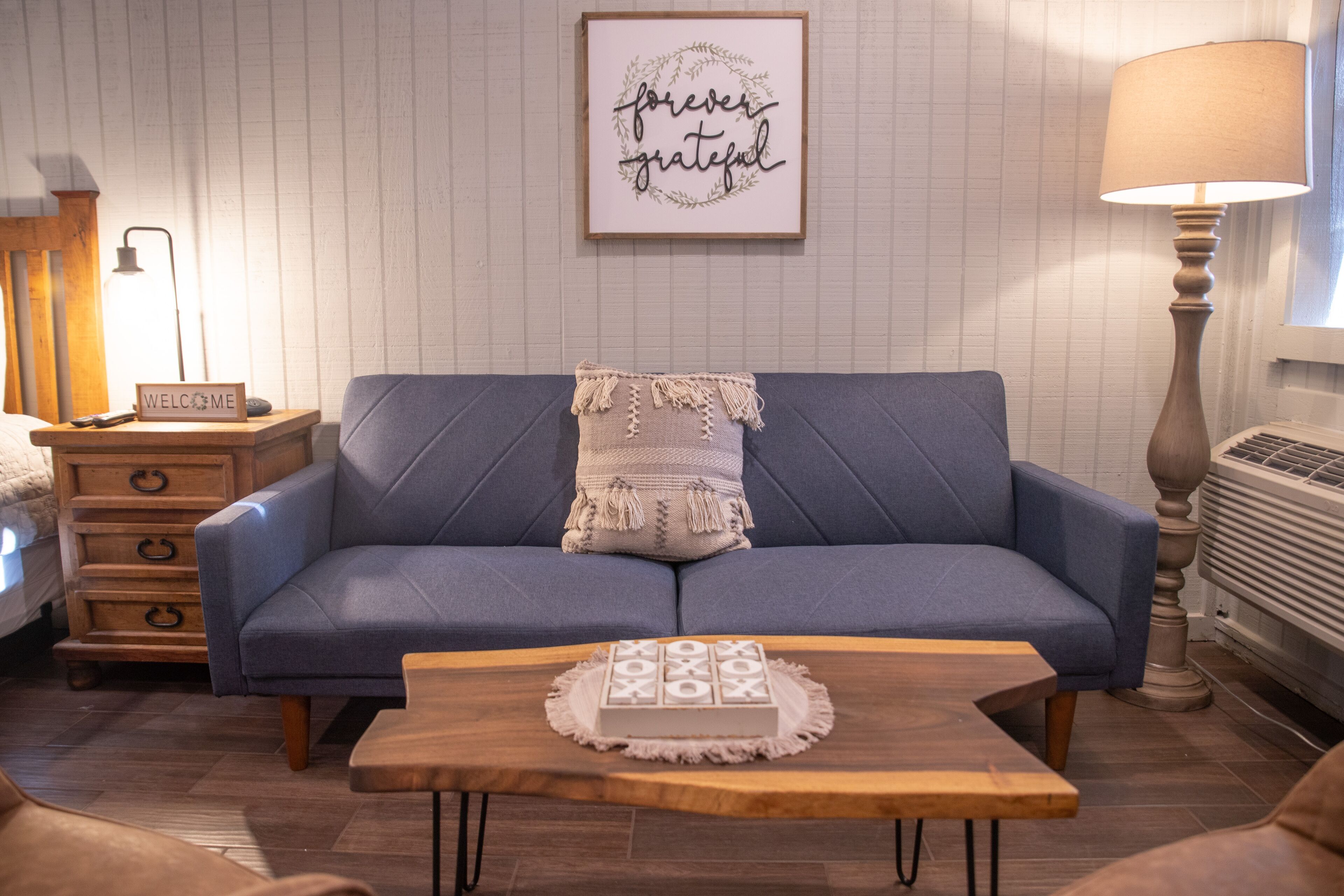 Una sala de estar con un sofá azul, una mesa de centro de madera con un objeto decorativo, una lámpara y un cartel enmarcado en la pared.
