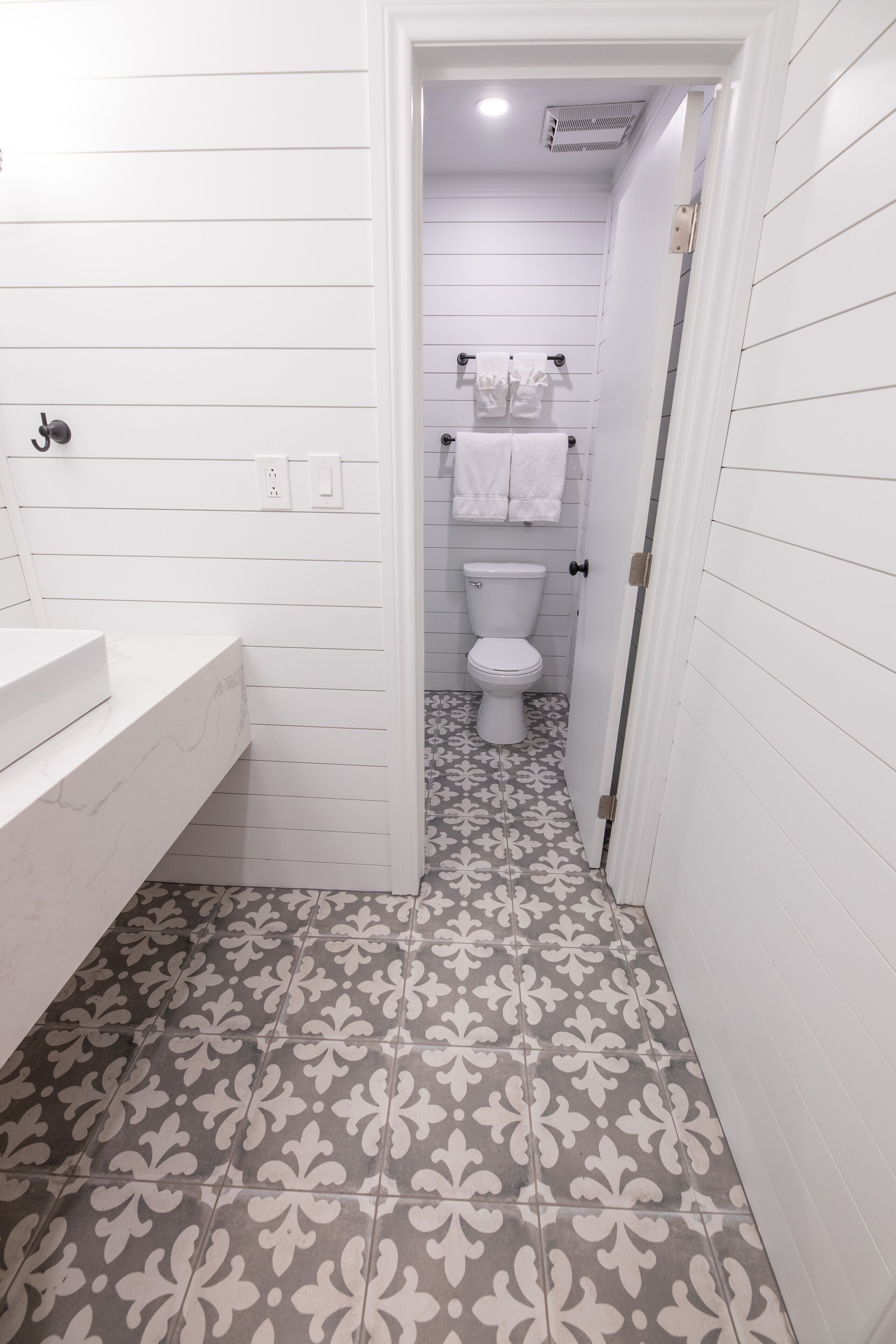 A narrow bathroom with white walls, a patterned floor, a white sink, and a toilet with a towel rack.