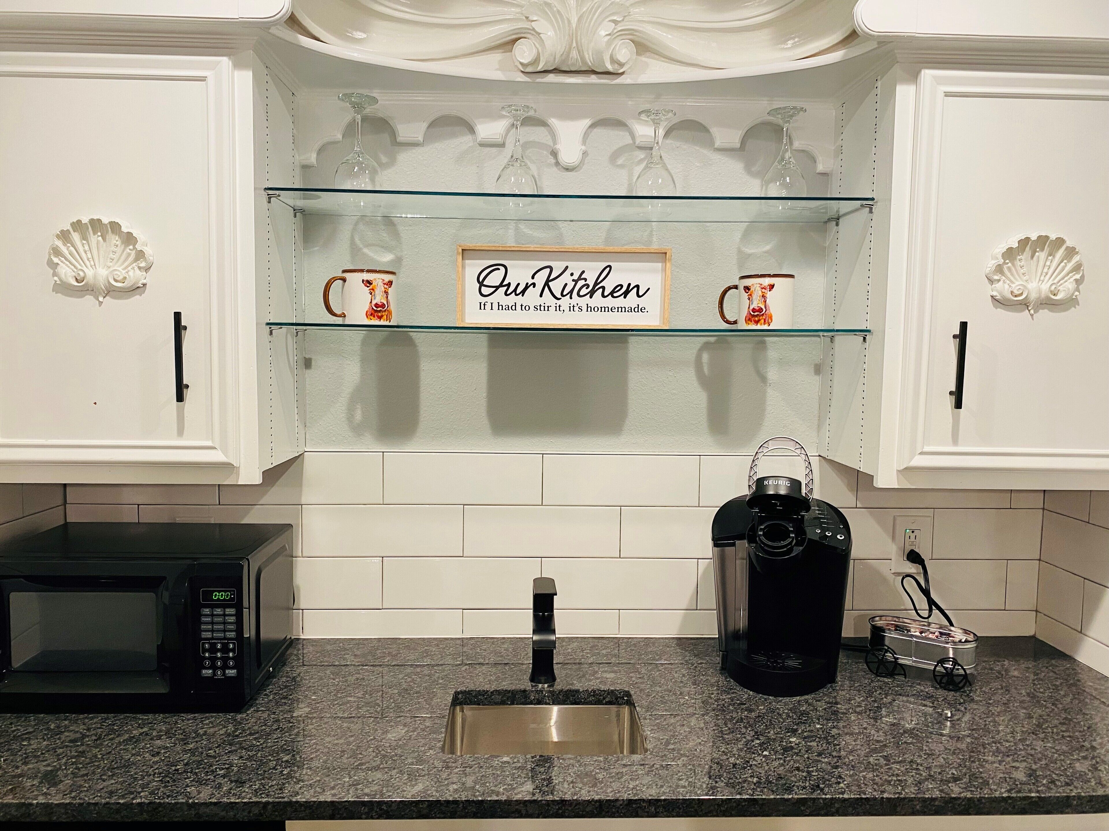 Uma cozinha com armários brancos, bancada de granito, cafeteira Keurig, micro-ondas e uma estante com objetos decorativos.