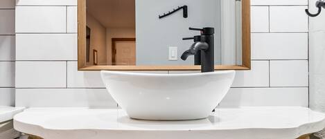 Un baño con lavamanos blanco, tocador de madera y un espejo con dos luces empotradas.