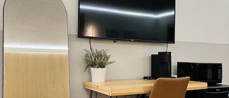 A modern room with a flat-screen TV mounted on the wall, a wooden desk with a chair, a small potted plant, and a black coffee machine.