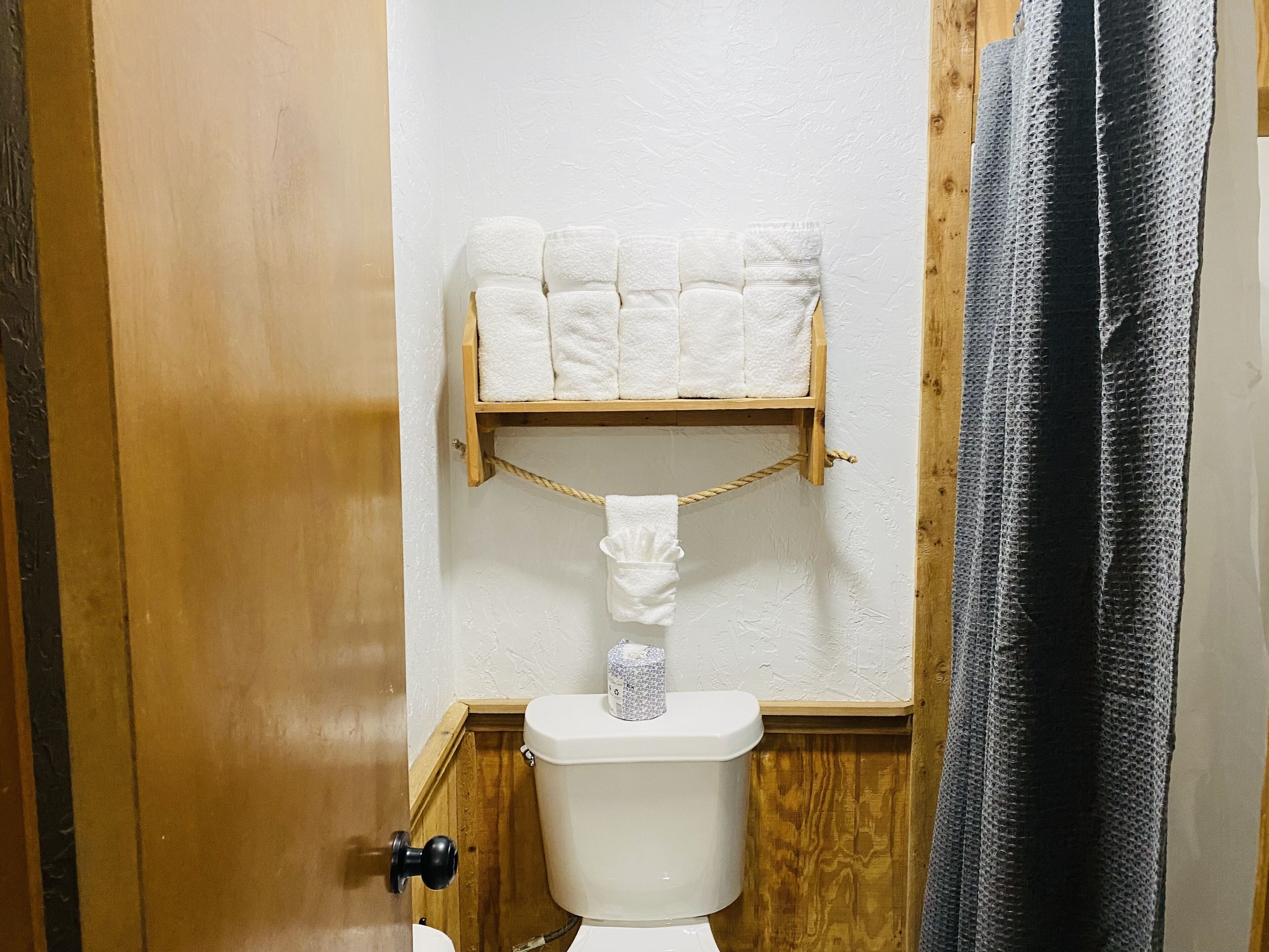 A bathroom with a wooden shelf holding white towels, a toilet, and a shower curtain.