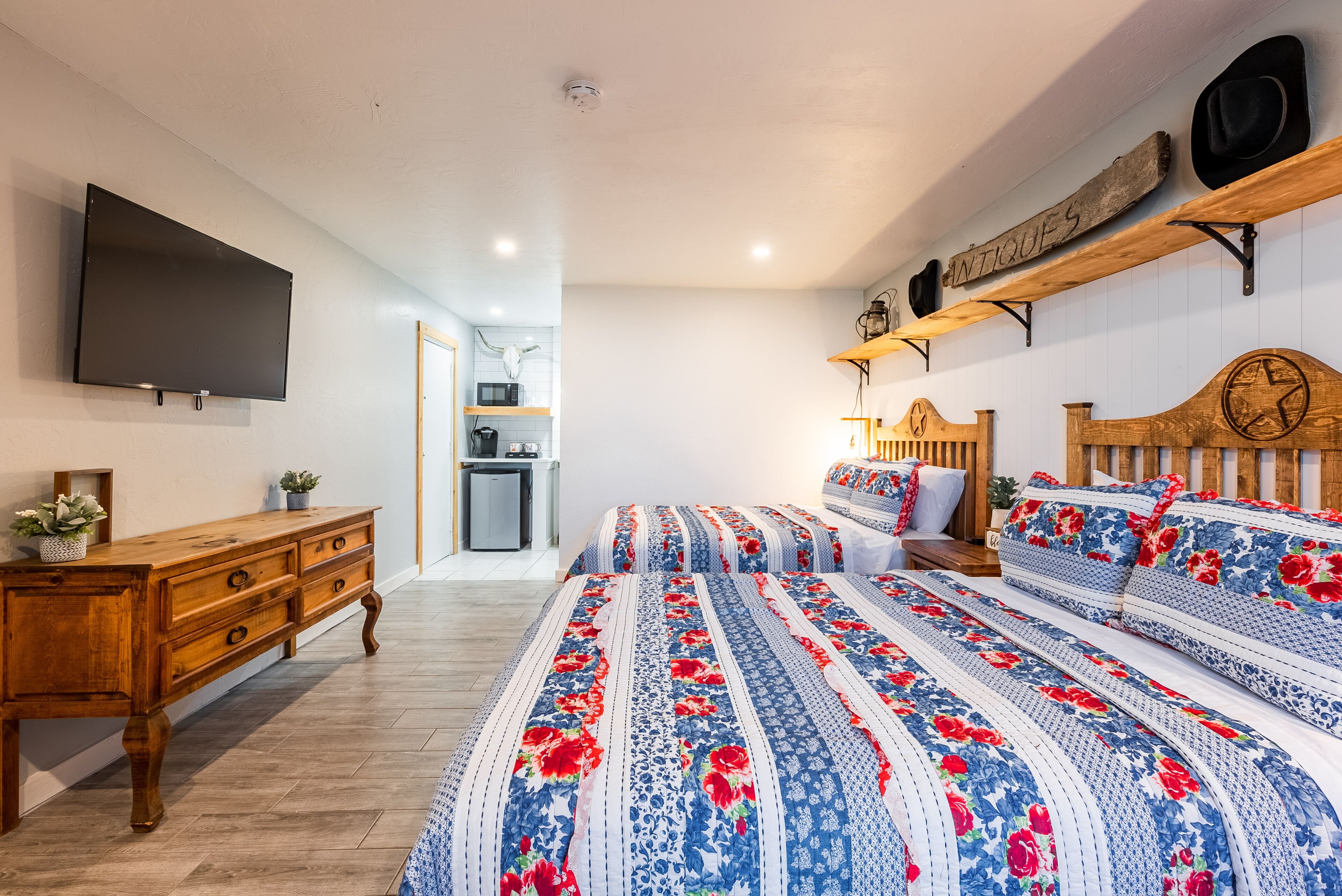 Un dormitorio con un aparador de madera, una cama con ropa de cama floral, un televisor montado en la pared y un estante con sombreros y un letrero que dice "ANTICQUES".