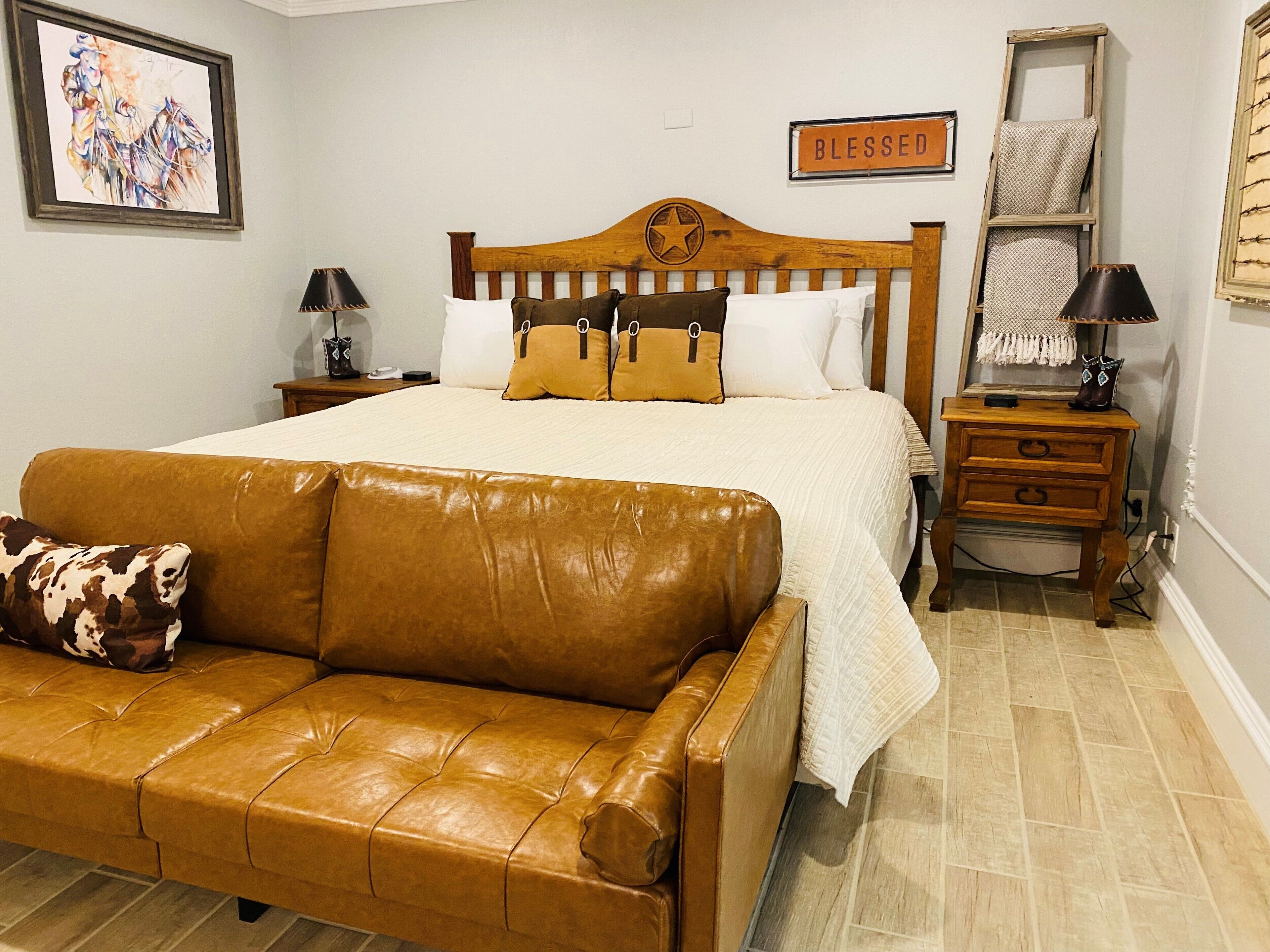 A bedroom with a large bed, a wooden headboard, a brown leather sofa, a nightstand, a lamp, and a framed picture on the wall.