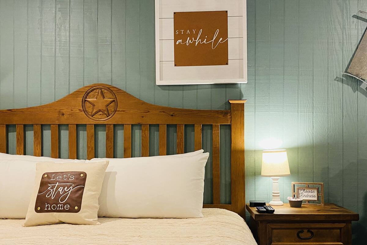 A bedroom with a wooden headboard, a bed with two pillows, a nightstand with a lamp, and a framed sign on the wall.