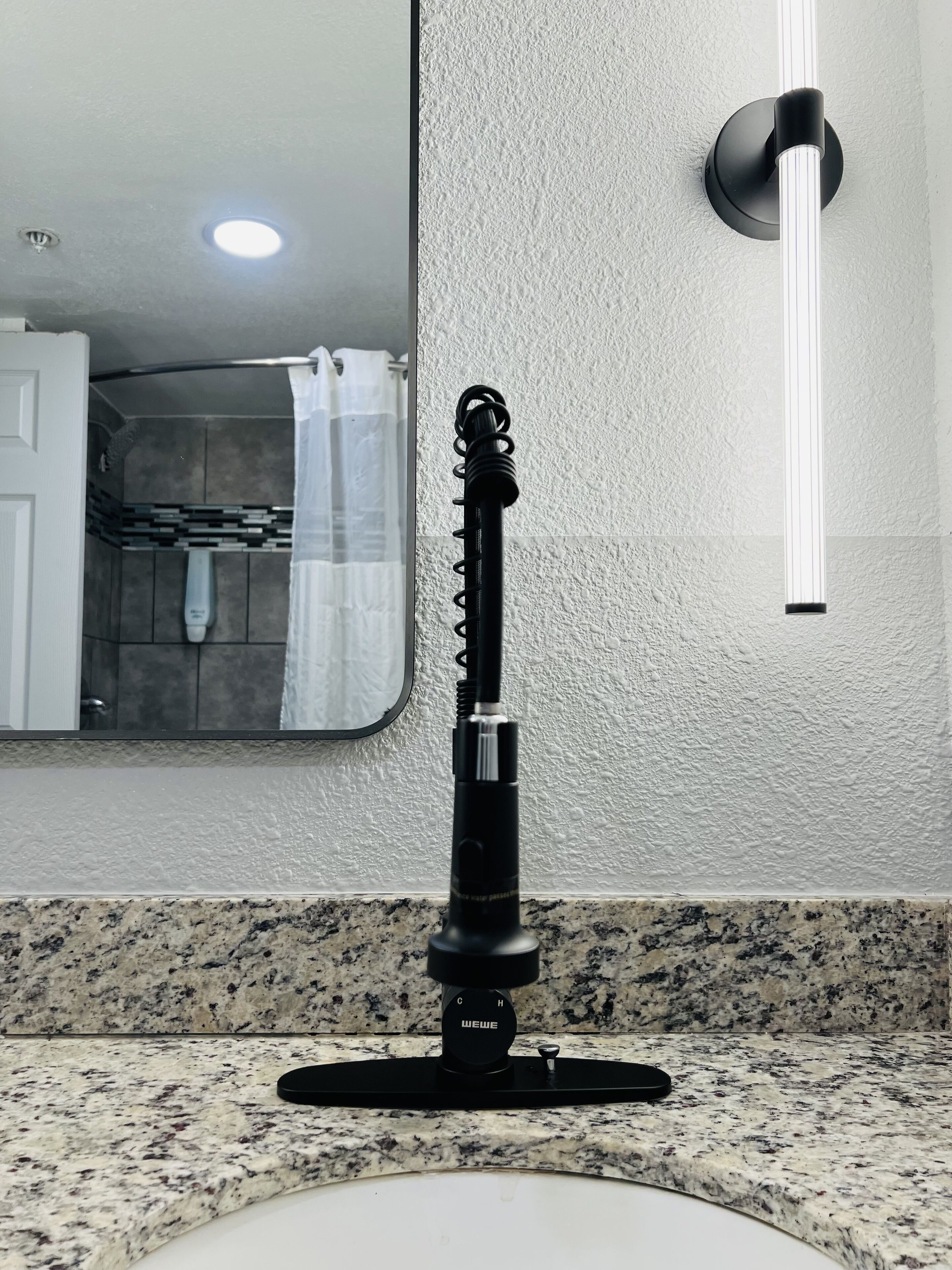A bathroom with a granite countertop, a black electric shaver on the sink, a mirror, and a wall-mounted light fixture.