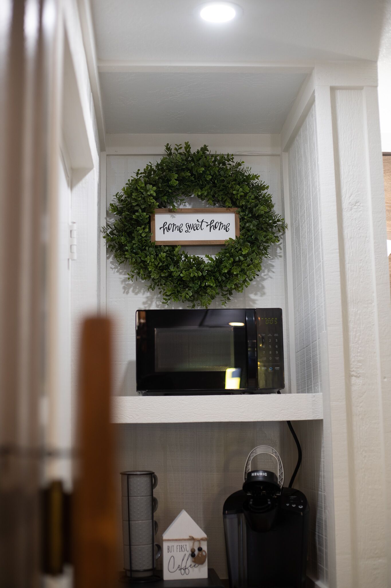 A kitchen with a microwave on a white shelf, a coffee maker, and a sign that says "home sweet home" above a green wreath.