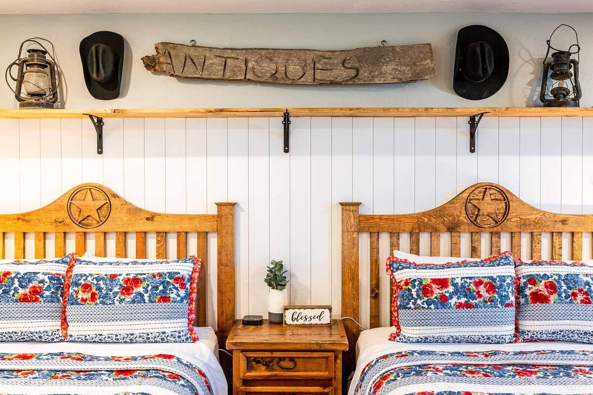 A room with two wooden beds, each with floral-patterned bedding and a star emblem on the headboard. A wooden sign above the beds reads "ANTIQUE."