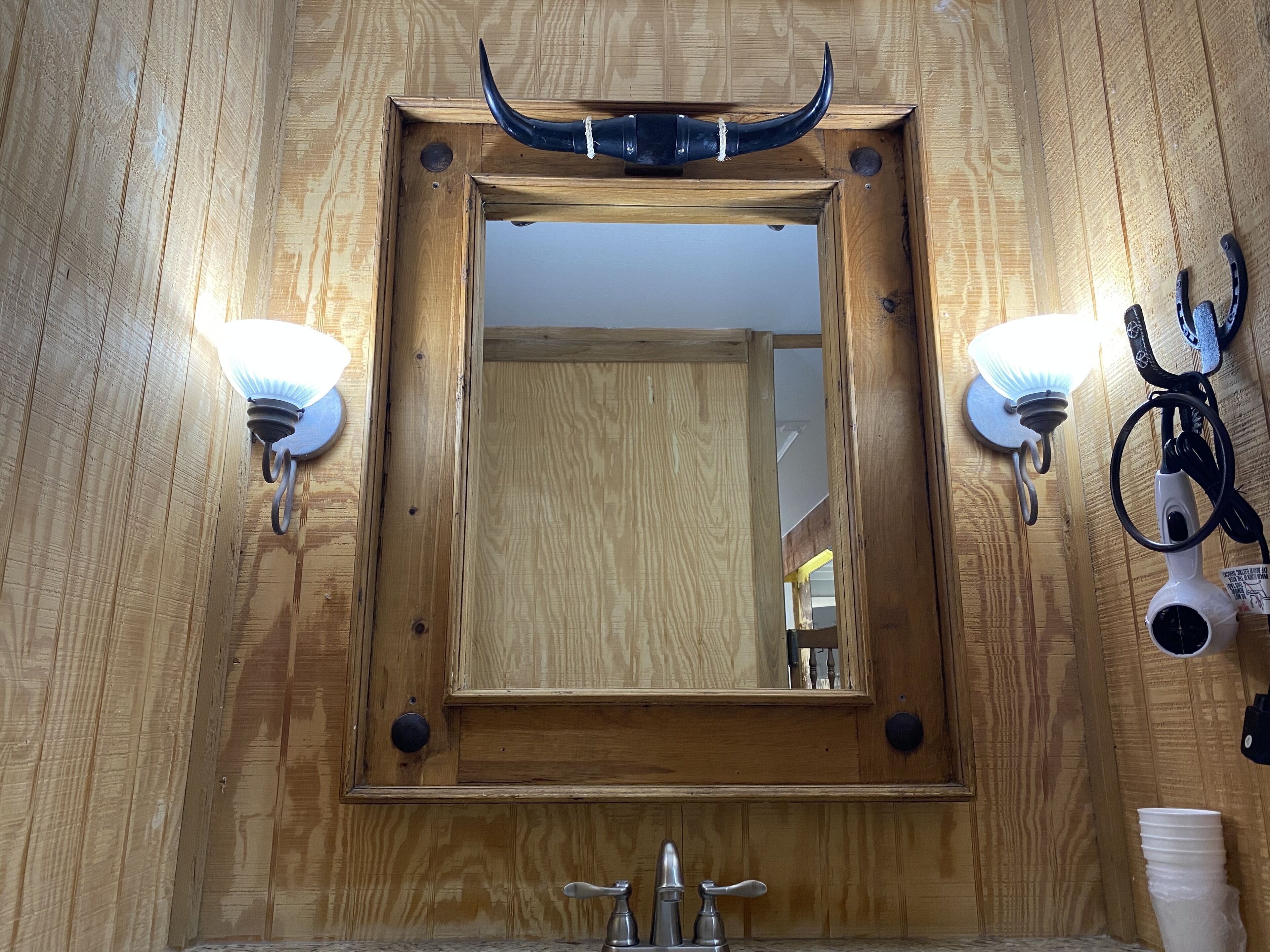 A wooden-framed mirror with two wall sconces, a sink with a faucet, and a horn above the mirror.