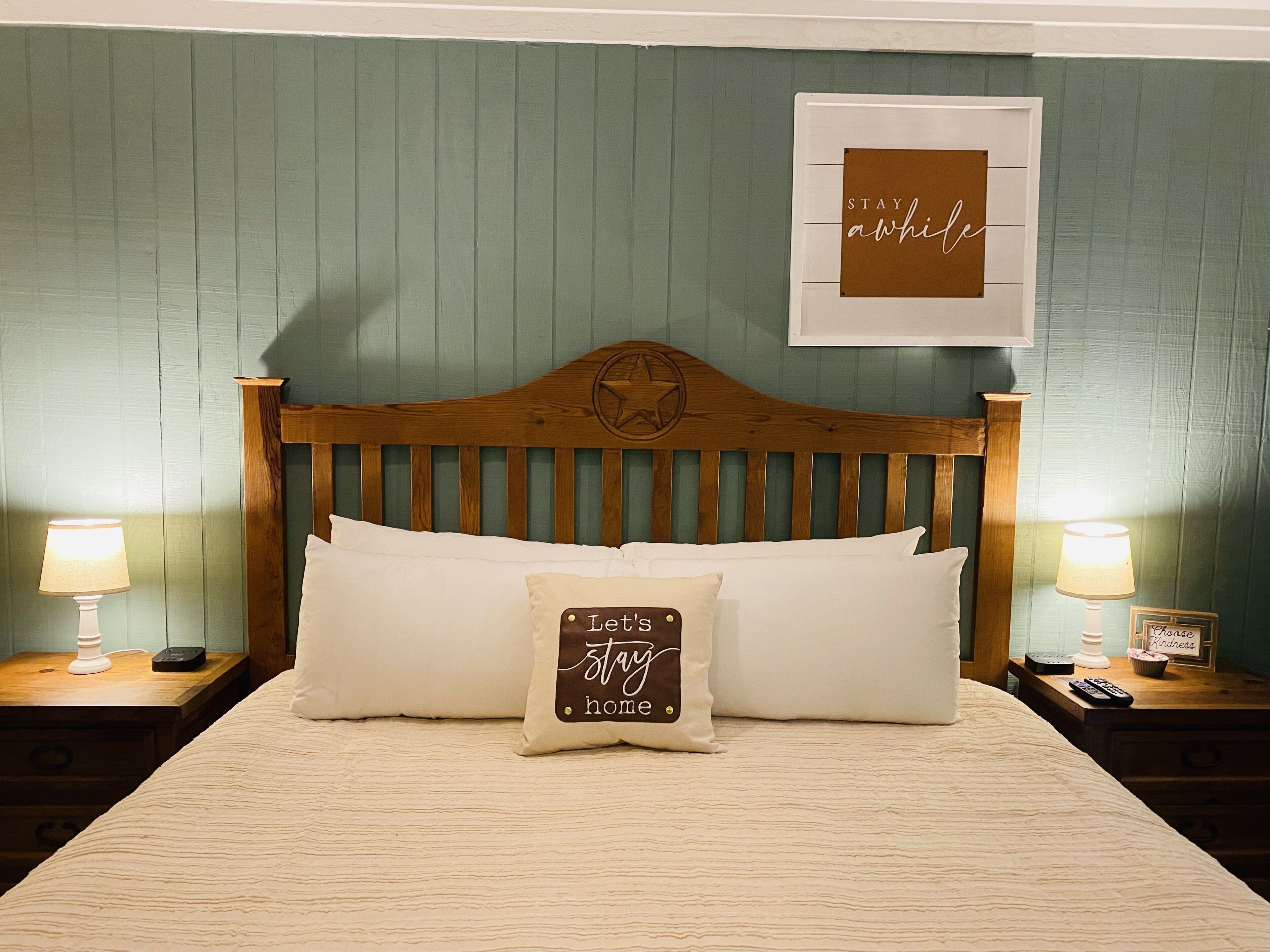 A bedroom with a wooden headboard, two bedside lamps, a framed sign on the wall, and a pillow with a message.