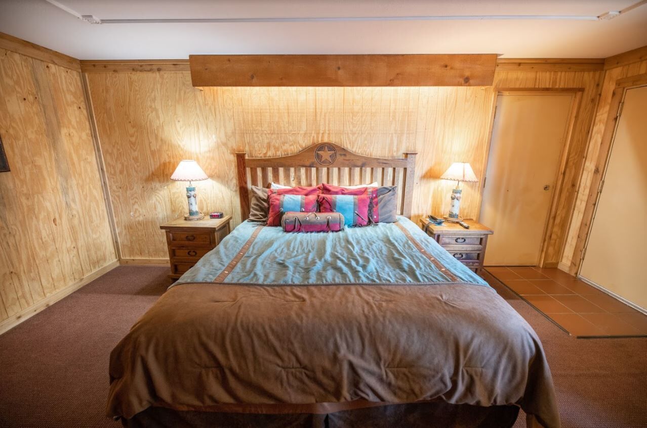 A bedroom with a wooden headboard, a bed with a blue and brown bedspread, two bedside tables with lamps, and a wooden wardrobe.