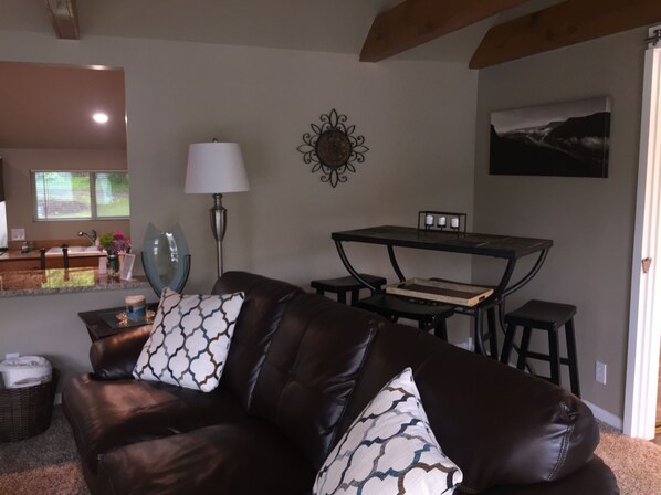A living room with a brown leather sofa, patterned cushions, a side table, a lamp, a wall decoration, and a dining area with a table and chairs. - Little house on the river (Chattanooga)